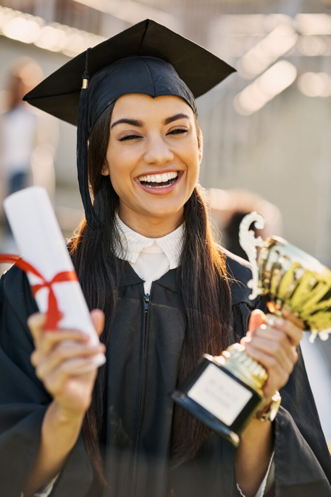 image shows a smiling woman in graduation attire, holding a rolled-up diploma tied with a red ribbon in one hand and a golden trophy in the other